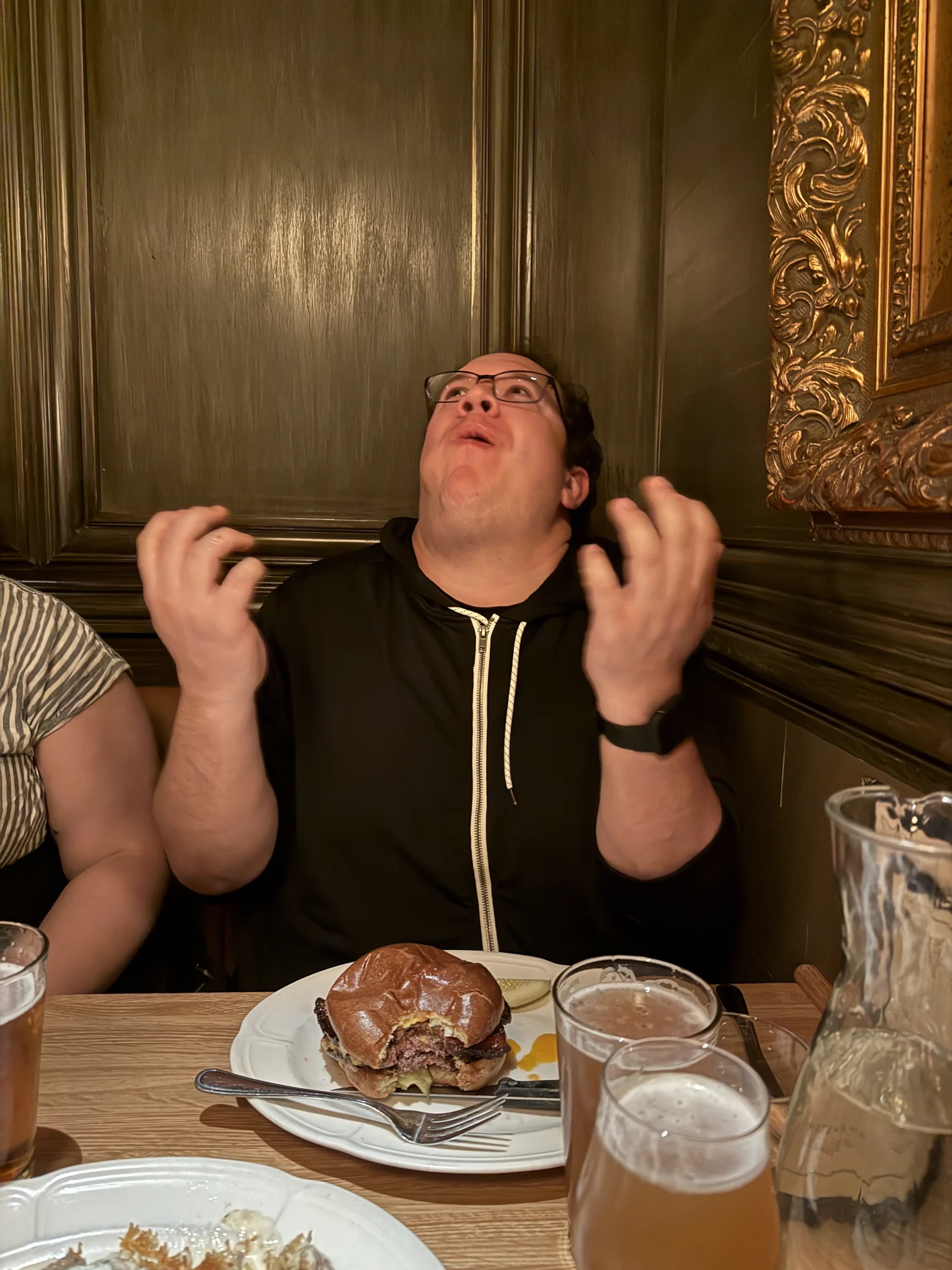 A delighted man at a restaurant looks upward with both hands raised; a burger and drinks sit on the table